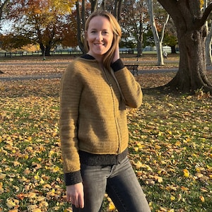 Könnte beinhalten: Eine Frau trägt einen braunen Strickpullover mit Reißverschluss und schwarzen Besätzen, graue Jeans und einen Hintergrund aus Herbstlaub.