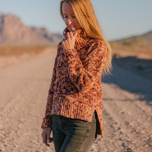 Könnte beinhalten: Eine Frau trägt einen mehrfarbigen, grob gestrickten Pullover mit Rollkragen und seitlichem Schlitz. Der Pullover ist eine Mischung aus Rosa, Orange und Gelb.