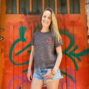 Könnte beinhalten: Eine Frau trägt ein braun-graues gestreiftes T-Shirt mit einer kleinen Tasche auf der linken Seite. Sie trägt Jeans-Shorts und hat langes blondes Haar. Sie steht vor einer leuchtend orangefarbenen Tür mit grünen und violetten Graffiti.