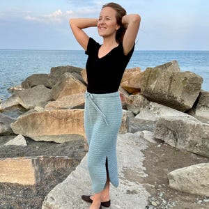 May include: A light blue, knee-length crochet skirt with a drawstring waist and a side slit. The skirt is paired with a black v-neck top. The background features a rocky shoreline and the ocean.