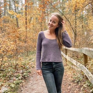May include: A woman wearing a purple long-sleeved sweater and dark wash jeans stands on a path in a forest. The trees have yellow and orange leaves.