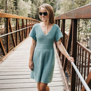 May include: A light teal crocheted dress with a V-neck and short sleeves. The dress has a fitted waist and a flared skirt. The woman is wearing sunglasses and standing on a wooden bridge with a metal railing. The background features trees and a natural setting.