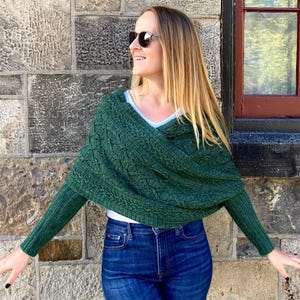 May include: A green knitted wrap with sleeves, featuring a textured cable knit design. The wrap is worn over a white top and blue jeans. The woman is wearing sunglasses and is standing in front of a stone wall.