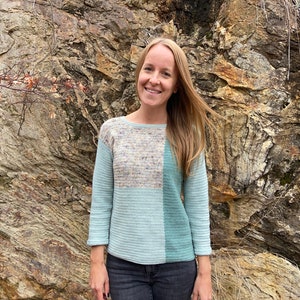 May include: A woman wearing a light blue, green and beige sweater with a colour block design. She is standing in front of a rocky wall.