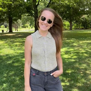 May include: A light green, sleeveless, button-down crochet top with a collar. The top is paired with dark gray denim shorts. The person is wearing round, gold-rimmed sunglasses. The background is a park with green grass and trees.