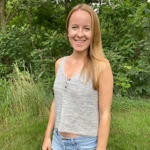 May include: A woman wearing a gray crochet tank top with a button closure and denim shorts. She is standing in a grassy area with trees in the background.