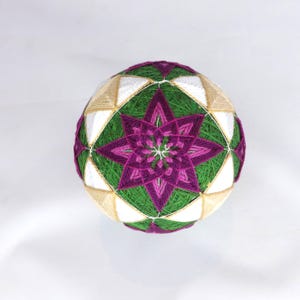 May include: A spherical temari ball featuring intricate embroidery with green, purple, magenta, white, and tan thread. The surface displays a geometric pattern of interconnected triangles and a central star-like motif created with layered stitching.