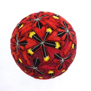 May include: A handcrafted, spherical Temari ball. The ball features a complex geometric design with red, black, and yellow thread. The intricate pattern is stitched over a soft core, creating a unique decorative item.