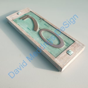 Op de afbeelding: Een houten huisnummerbord met een teal groene achtergrond en een koperkleurig nummer "70" erop. Het bord heeft een verweerde witte verfbehandeling.