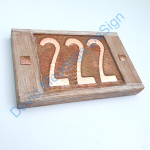 Op de afbeelding: Een houten plaquette met een koperen achtergrond en het nummer 222 in witte letters. De plaquette heeft een verweerde witte verfbehandeling en is versierd met koperen klinknagels.