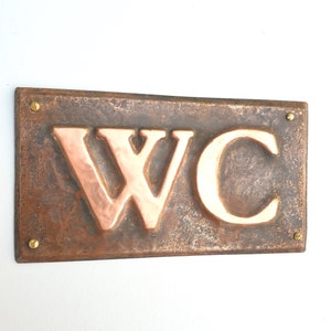 May include: A rectangular copper sign with the letters "WC" embossed in copper. The sign has a rustic, aged look and is secured with brass screws.