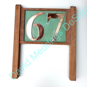 May include: A house number sign featuring the digits 67 in polished copper mounted on a rectangular green panel. The panel is framed by a wooden border with two vertical wooden posts extending downward from the sides of the frame.