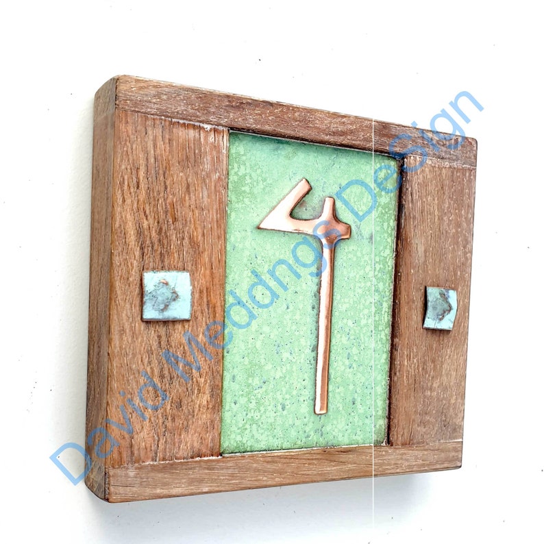 Op de afbeelding: Een koperkleurig metalen nummer vier is gemonteerd op een groene tegel met een houten frame. De tegel heeft een gespikkelde groene glazuur. Het frame is gemaakt van donker hout met een lichte houten rand.