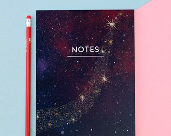 Op de afbeelding: Een notitieboek met een donkerblauw, rood en goud hemels ontwerp. De cover heeft het woord "NOTES" in het wit, met een sterrenbeeldpatroon. Een rode potlood staat naast het notitieboek.