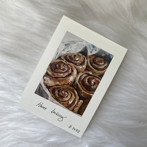 May include: A painting of a plate of freshly baked cinnamon rolls. The rolls are a light brown colour and have a swirl pattern. The painting is titled "Home Baking" and dated 8-2022.