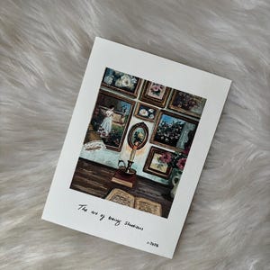 May include: A white card featuring a painting of a room with framed pictures, a candle, and an open book. The text "The art of being studious" is handwritten on the card. The card is on a fluffy, white surface.