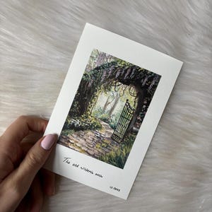 Puede incluir: Una pintura a la acuarela de un camino de piedra que conduce a una puerta de hierro forjado bajo un arco de glicinas. La pintura está sobre una tarjeta blanca con el texto "The old wisteria arch" y la fecha "12.2023".