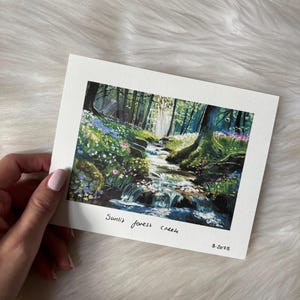 May include: A hand holding a watercolor painting of a forest scene. The artwork depicts a sunlit forest creek with lush greenery, wildflowers, and flowing water. The text "Sunlit forest creek" and the date "8.2025" are written on the painting.