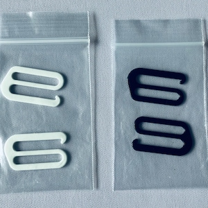 May include: Two sets of bra strap hooks, one white and one black, each in a clear plastic bag. The hooks are made of a sturdy material and are designed to attach to bra straps. The bags are sealed with a zipper closure.