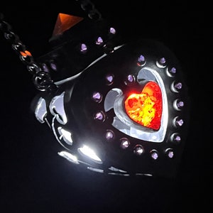 May include: A black metal heart-shaped pendant with a smaller red heart inside. The pendant is adorned with small purple rhinestones around the edges.