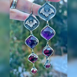 May include: Silver-toned dangle earrings featuring a bat emblem at the top, followed by a large, faceted purple gemstone, a smaller red gemstone, and a tiny red gemstone at the bottom. The earrings are displayed against a blurred green background.
