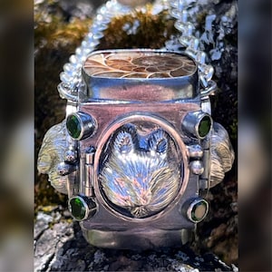 May include: A silver pendant necklace with a wolf head design, a fossilized stone, and green gemstones. The pendant is attached to a silver chain.