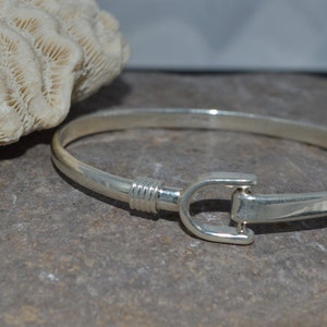 Könnte beinhalten: Ein silbernes Armreif-Armband mit einer einfachen, polierten Oberfläche. Das Armband verfügt über einen einzigartigen Verschluss, der einem Haken ähnelt.