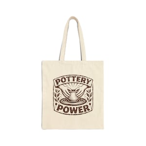May include: Cream-colored tote bag with a brown graphic. The graphic features hands shaping clay on a pottery wheel, with the words "POTTERY POWER" in a vintage style. The bag has two handles for carrying.