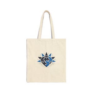 May include: A cream-coloured tote bag featuring a blue and navy heart-shaped jigsaw puzzle design. The design is surrounded by a starburst pattern. The bag has two handles and is made of a canvas-like material.