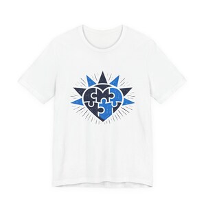 Peut inclure: T-shirt blanc avec un motif en forme de cœur bleu et noir composé de pièces de puzzle. Le cœur est entouré d'un motif d'éclat. Le t-shirt est fait d'une matière douce.