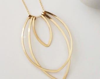 May include: A gold-coloured necklace with a fine chain and a pendant comprising three overlapping, leaf-shaped elements. The pendant has a minimalist, modern design with clean lines and a polished finish. The necklace is displayed on a white surface.