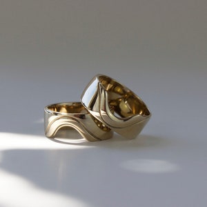 May include: Two gold-colored rings with a unique wave design. The rings are stacked on top of each other, with one slightly angled. The rings have a polished, reflective surface, and are set against a white background.