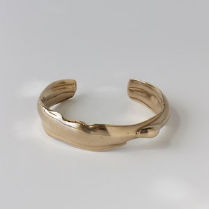 May include: A gold-colored cuff bracelet with a textured, organic design. The open-ended bracelet has a flowing, irregular shape, suggesting a natural or handcrafted aesthetic. The metal has a polished finish.