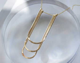 May include: A gold necklace with a geometric design. The necklace features three curved, U-shaped pendants connected by a delicate chain. The pendants are arranged in a layered style, creating a modern and minimalist aesthetic. The necklace is displayed on a clear, reflective surface.
