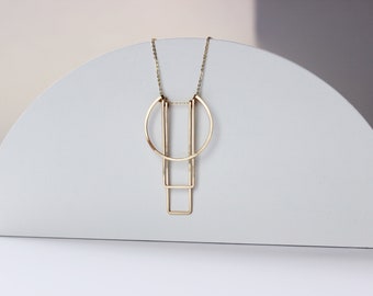 May include: A gold-coloured necklace with a geometric design. The necklace features a half-circle, two vertical bars, and a square shape. The chain is delicate, and the pendant is suspended against a white background.