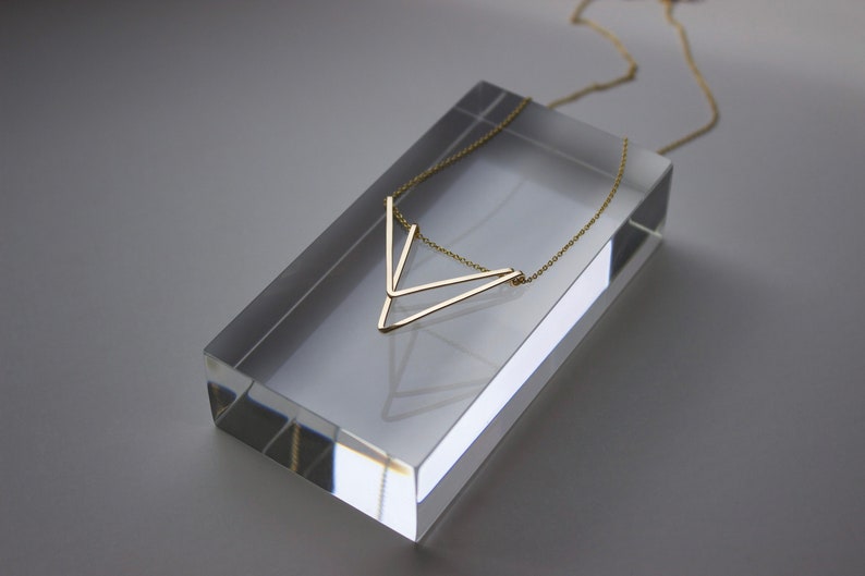 May include: A gold-colored necklace with a geometric V-shaped pendant rests on a clear acrylic block. The necklace has a delicate chain and a minimalist design, suitable for everyday wear.