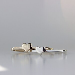 Puede incluir: Dos anillos simples en forma de coraz&oacute;n, uno dorado y otro plateado, est&aacute;n apilados uno encima del otro. Los anillos est&aacute;n sobre una superficie blanca.