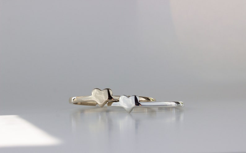 Puede incluir: Dos anillos simples en forma de coraz&oacute;n, uno dorado y otro plateado, est&aacute;n apilados uno encima del otro. Los anillos est&aacute;n sobre una superficie blanca.