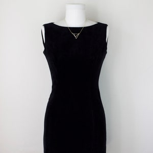 May include: A black sleeveless dress with a boat neckline displayed on a white mannequin. A delicate gold-colored necklace with a geometric triangle pendant adorns the neckline. The dress has a fitted silhouette.