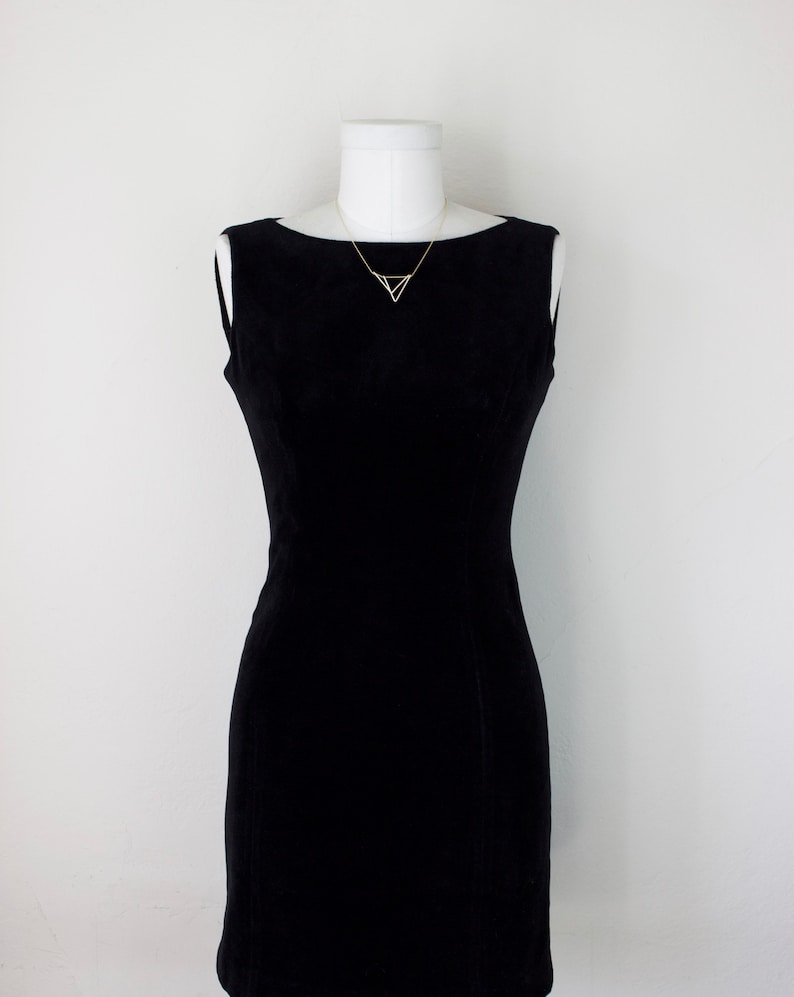 May include: A black sleeveless dress with a boat neckline displayed on a white mannequin. A delicate gold-colored necklace with a geometric triangle pendant adorns the neckline. The dress has a fitted silhouette.