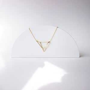 May include: A delicate gold-colored necklace with a geometric triangle pendant. The necklace is displayed on a white, semi-circular stand against a white background. The pendant's design is minimalist and modern.