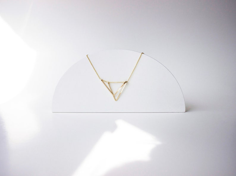 May include: A delicate gold-colored necklace with a geometric triangle pendant. The necklace is displayed on a white, semi-circular stand against a white background. The pendant's design is minimalist and modern.