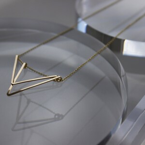 May include: A delicate gold-colored necklace with a geometric V-shaped pendant. The pendant is composed of thin, straight lines that form a modern, minimalist design. The necklace is displayed on a reflective surface.