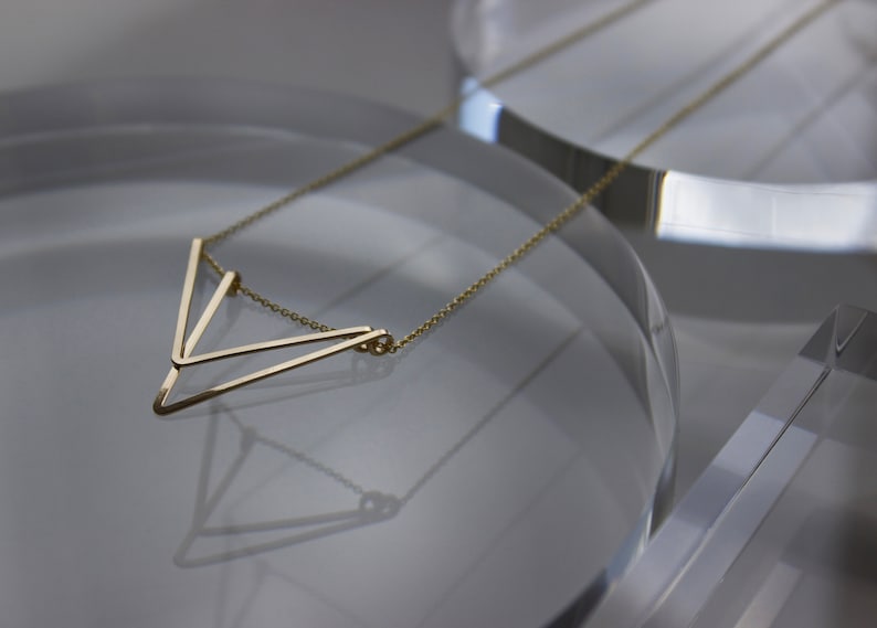 May include: A delicate gold-colored necklace with a geometric V-shaped pendant. The pendant is composed of thin, straight lines that form a modern, minimalist design. The necklace is displayed on a reflective surface.