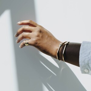 Puede incluir: Una mano con un anillo de oro y dos pulseras de oro. La mano se extiende hacia una pared blanca.