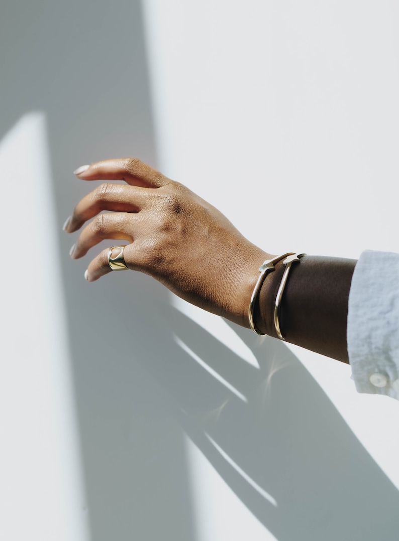 Puede incluir: Una mano con un anillo de oro y dos pulseras de oro. La mano se extiende hacia una pared blanca.