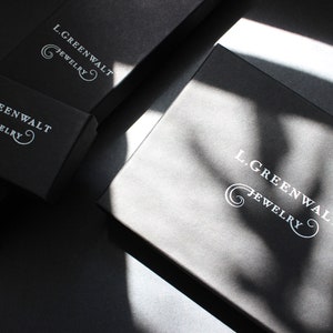 May include: Several black jewelry boxes are arranged on a dark surface. The boxes have the text "L. GREENWALT JEWELRY" printed in white. The boxes are in various sizes and shapes, with shadows cast across them.