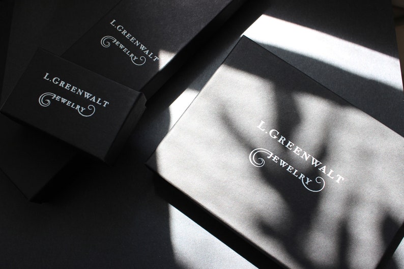 May include: Several black jewelry boxes are arranged on a dark surface. The boxes have the text "L. GREENWALT JEWELRY" printed in white. The boxes are in various sizes and shapes, with shadows cast across them.