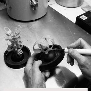 May include: A grayscale image shows a person working on a wax tree for lost wax casting. Two wax tree models sit on black bases. The person is using a pen-like tool. Industrial equipment is in the background.