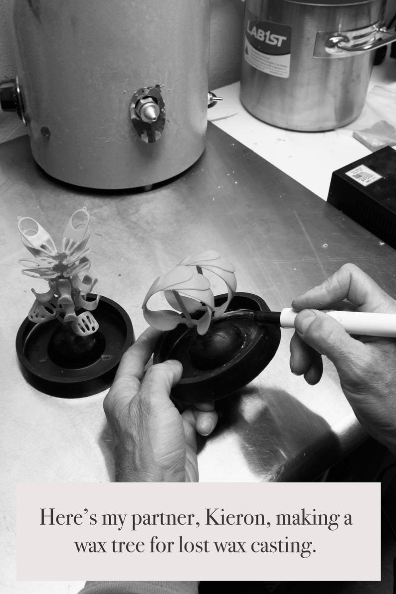 May include: A grayscale image shows a person working on a wax tree for lost wax casting. Two wax tree models sit on black bases. The person is using a pen-like tool. Industrial equipment is in the background.